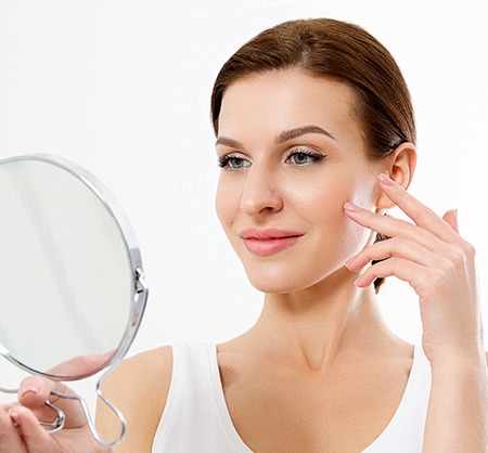 Woman applying skincare product while holding mirror, demonstrating beauty routine.