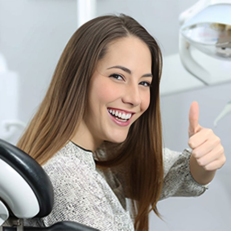 Young woman smiling and giving thumbs up in dental office setting with equipment visible.